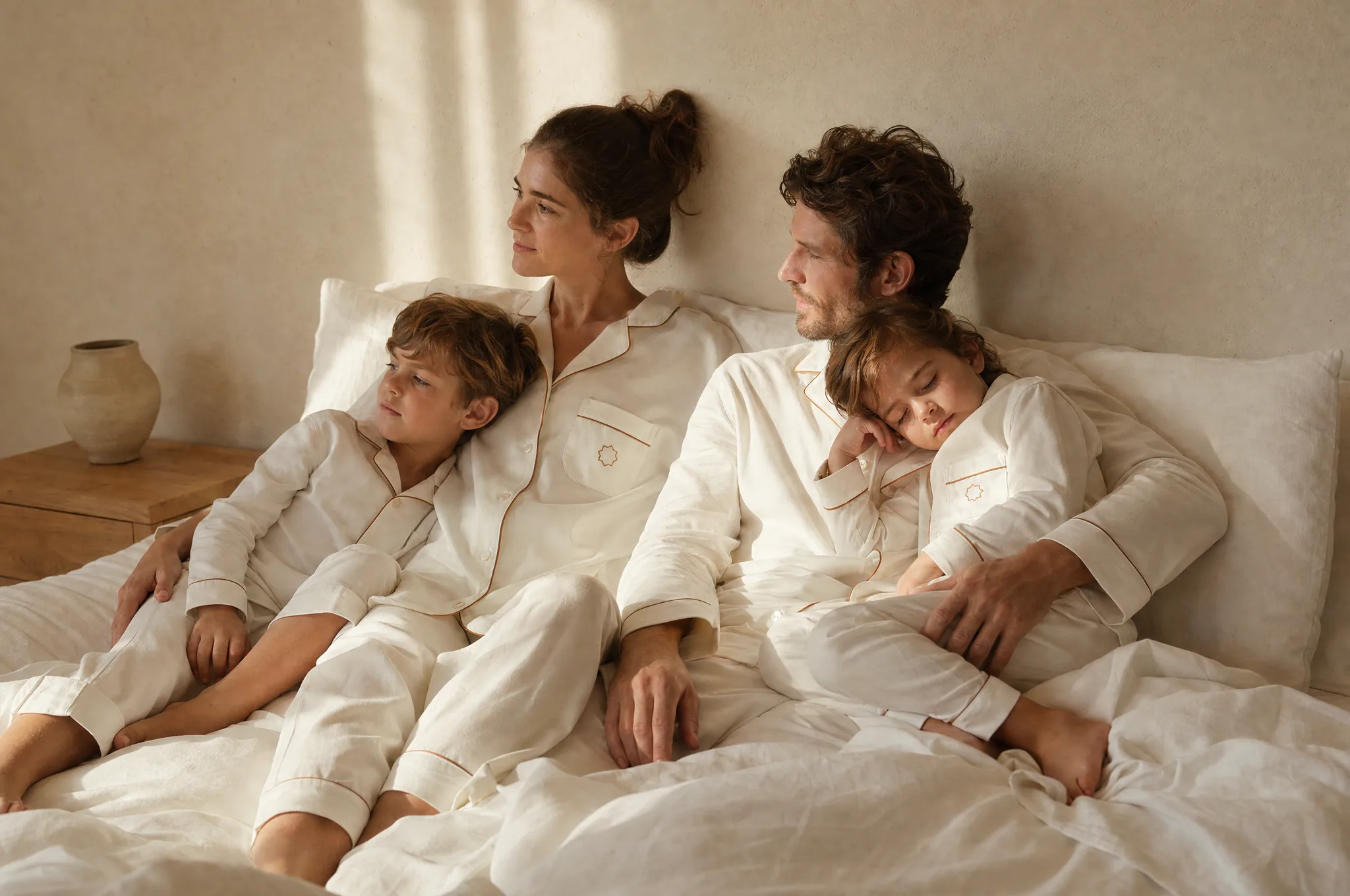 A family of four resting together on ivory linen in matching pajamas, morning light on a plaster wall