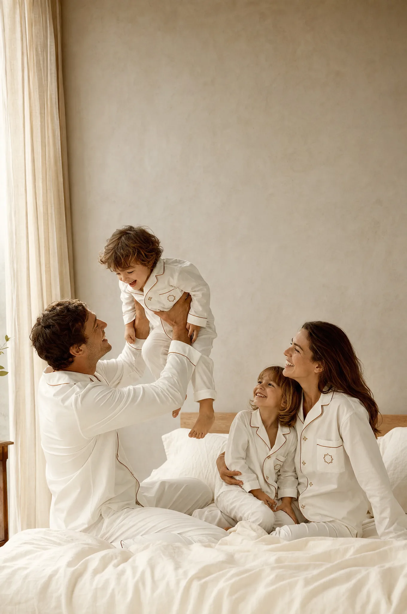 A father lifts his laughing toddler while mother and daughter watch from the bed, all in ivory pajamas with ginger piping